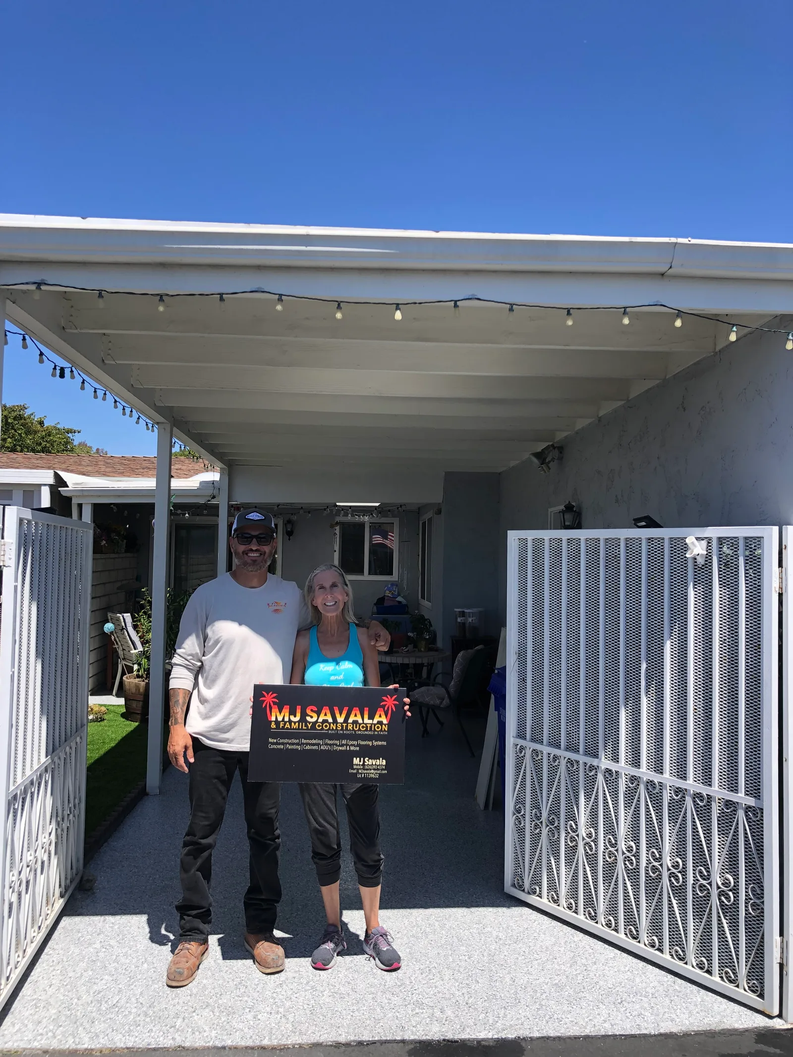 Two people standing on a newly coated gray speckled floor under a covered patio with open metal gates, one holding a company sign.