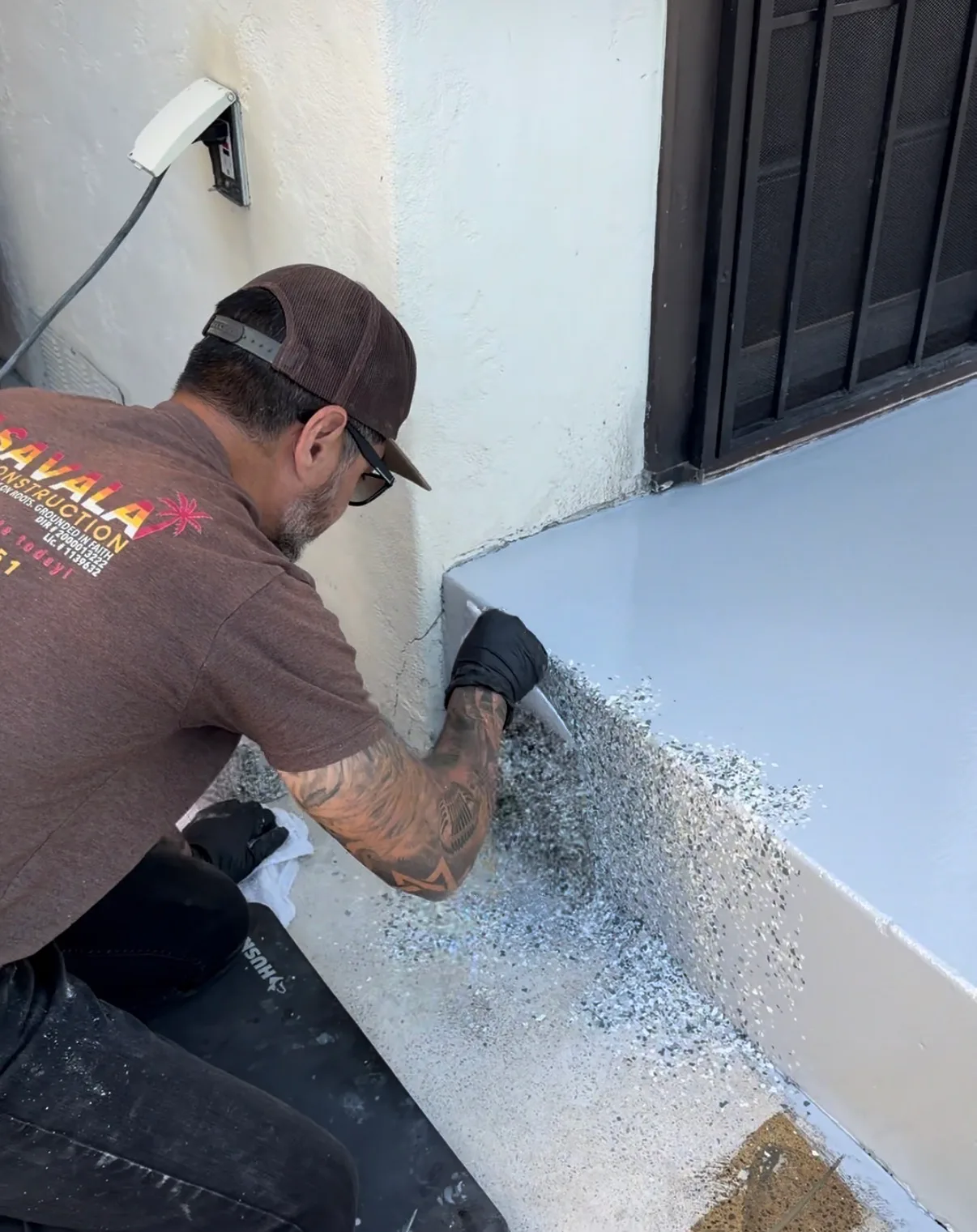 A person wearing a cap, glasses, and black gloves kneels while broadcasting fine gray flakes onto a freshly coated white surface near a wall and doorway.